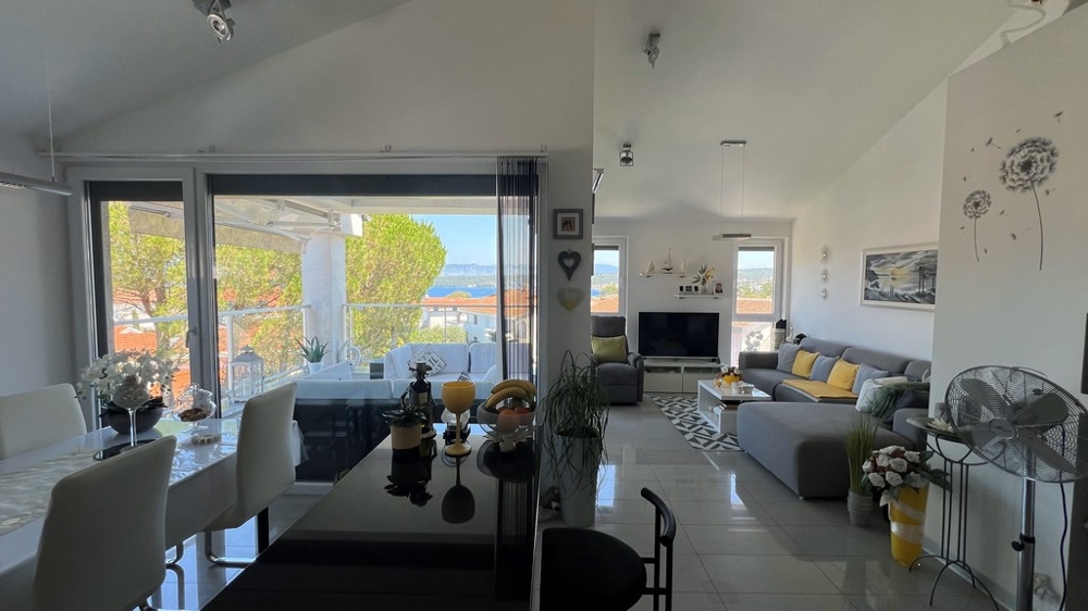 Luminoso soggiorno con divano grigio, zona pranzo e ampia vetrata verso la terrazza con vista mare.