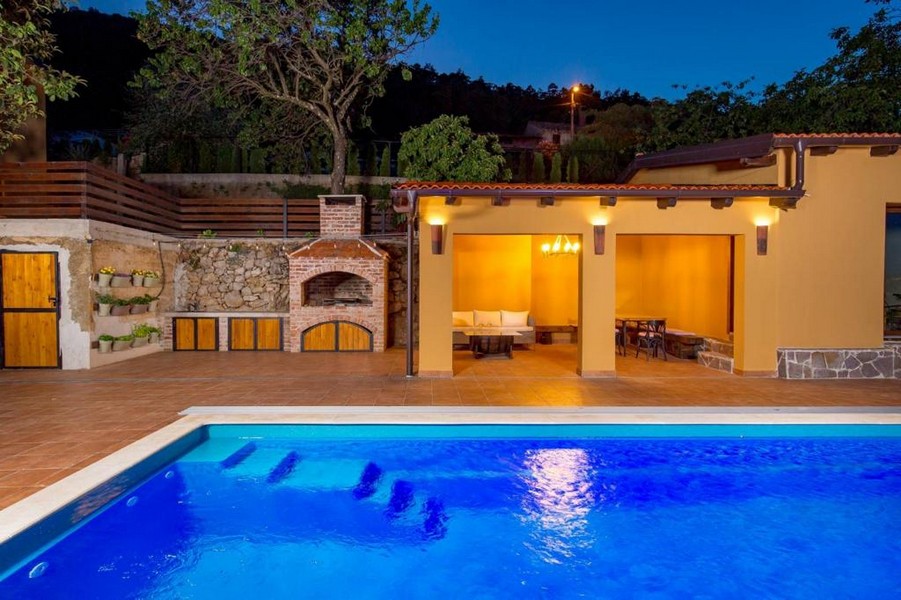 Piscina illuminata davanti alla cucina estiva con camino in muratura