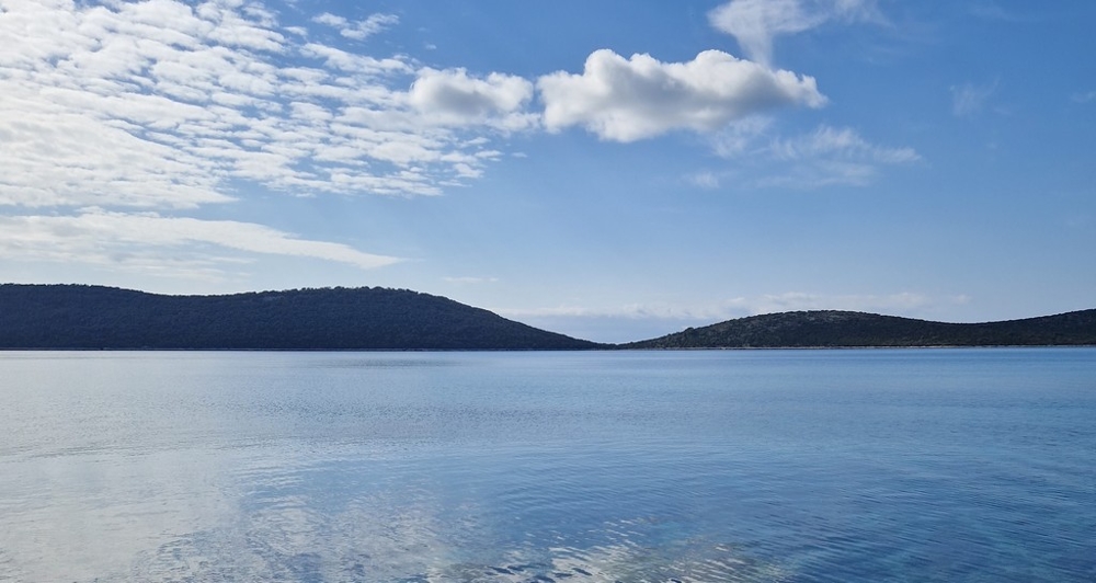 Specchio d'acqua tranquillo davanti a colline boscose sotto un cielo di nuvolette