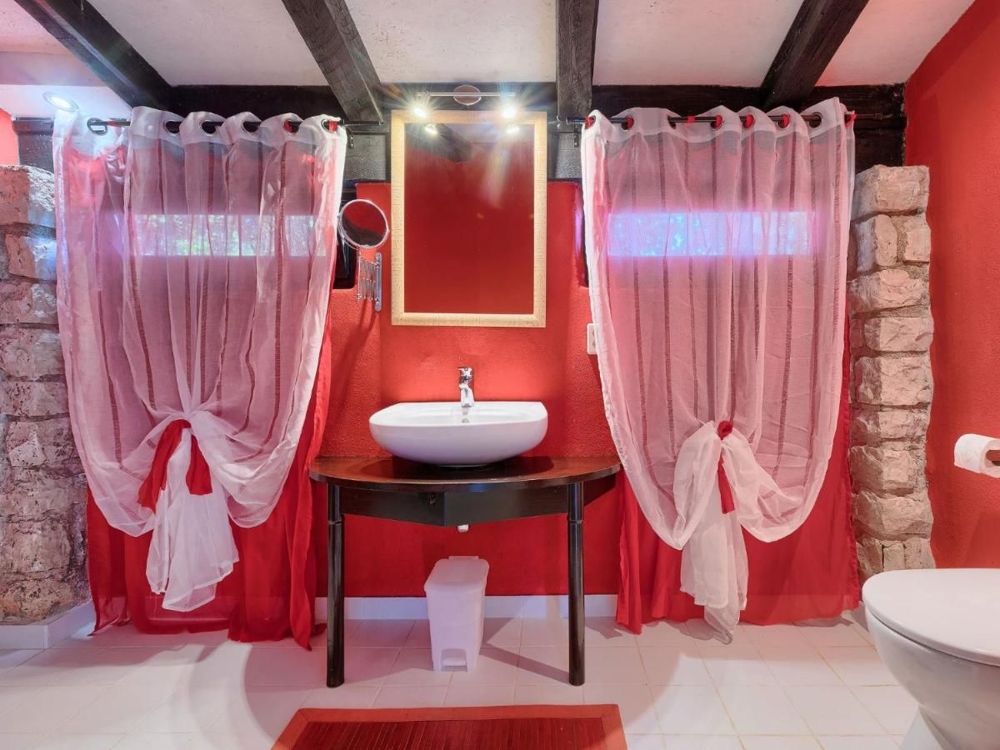 Bagno di un immobile in Croazia con pareti rosse, sanitari bianchi, un lavabo su un tavolo rustico e tende rosse trasparenti alle finestre