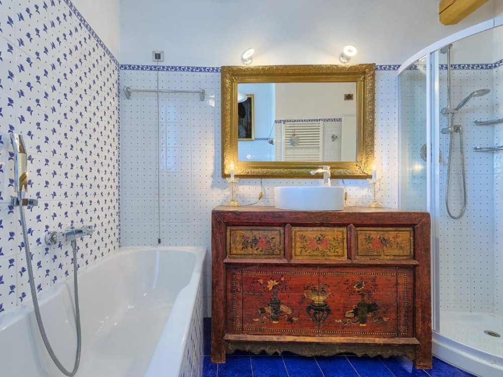 Un bagno in un immobile croato, decorato con piastrelle blu e bianche, una vasca, una zona doccia e un armadio antico e colorato sotto uno specchio dorato