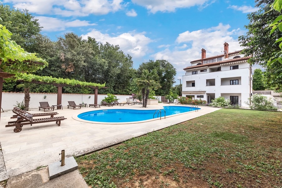 Una moderna area piscina con sdraio e una grande casa bianca sullo sfondo, circondata da alberi