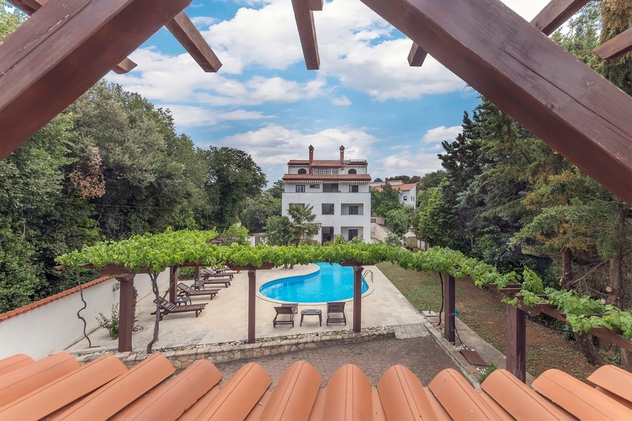 Una vista da una pergola sull'area piscina e sulla casa plurifamiliare bianca, circondata da vegetazione rigogliosa