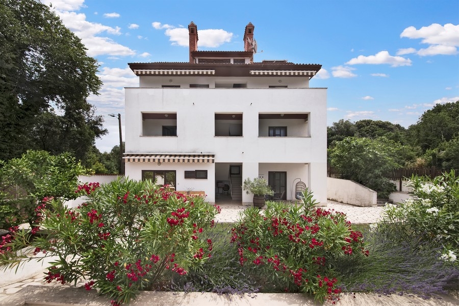La vista frontale della casa plurifamiliare bianca, circondata da cespugli in fiore e da un cielo azzurro.