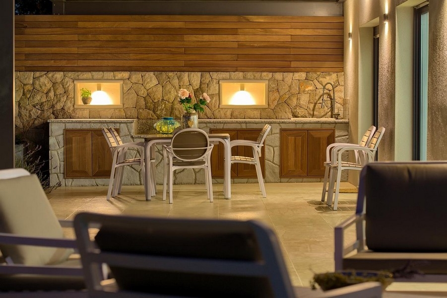Zona pranzo coperta all'aperto con cucina in pietra e armadi in legno, illuminata con stile