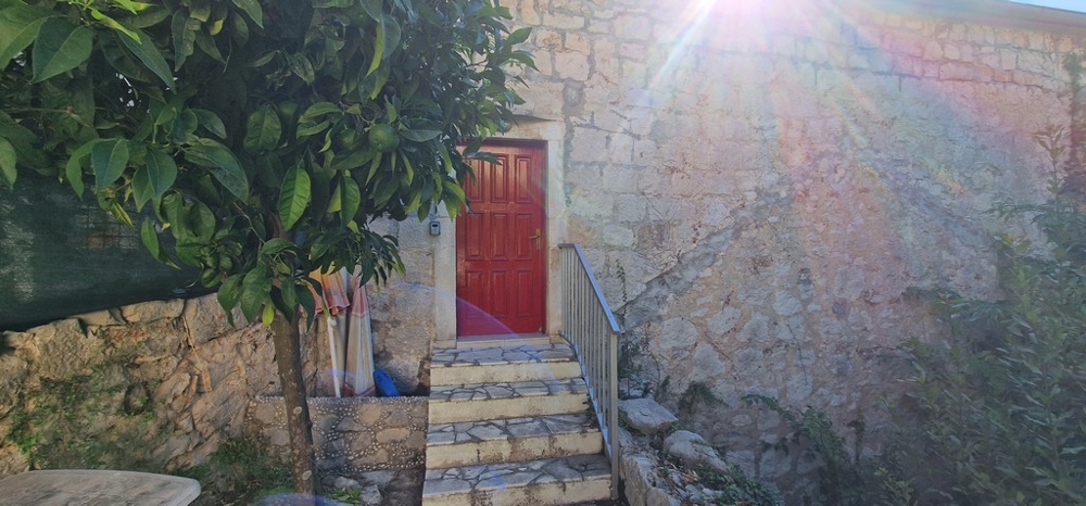 Una piccola area esterna con una scala in pietra che porta a una porta in legno rossa. La parete della casa è in pietra naturale e il sole splende nel cielo.