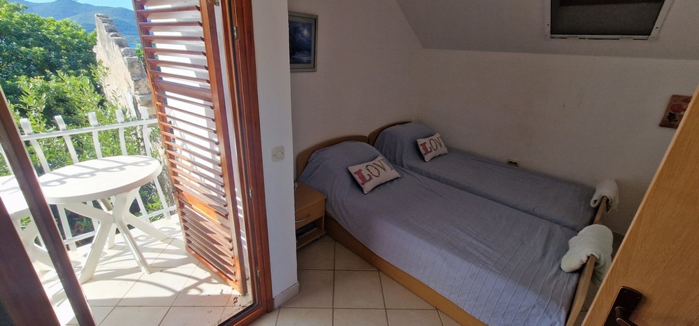 Una camera da letto con due letti singoli coperti da coperte blu. C'è un piccolo balcone con un tavolo rotondo e sedie e vista sul verde.