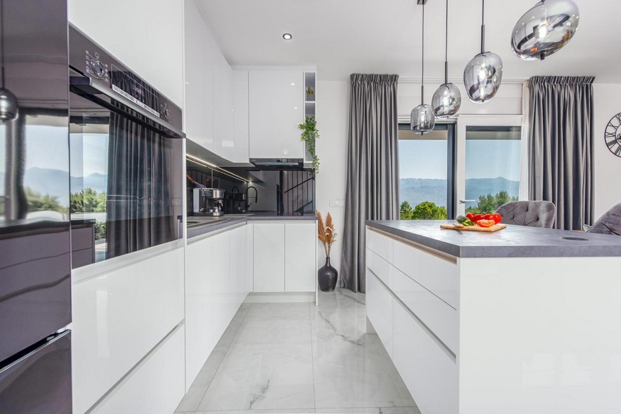 Cucina bianca moderna con isola, lampade a sospensione e vista sulla natura attraverso grandi finestre.